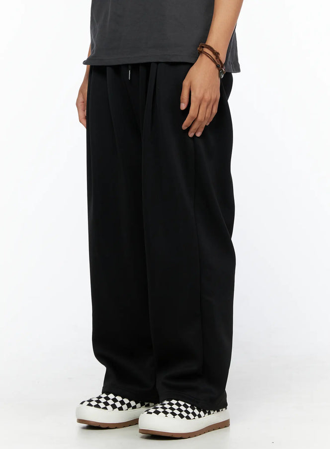 Men's Triple Tuck Sweatpants IL520 Black angled side view on model showing wide-leg drape, pleated front, and drawstring elastic waistband with checkered slip-on sneakers