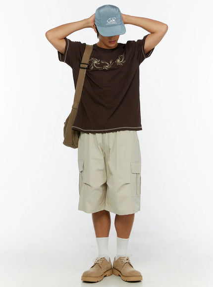 Men's Utility Cargo Shorts IL513 Beige full-length front view on model with hands raised behind head, styled with brown tee and crossbody bag