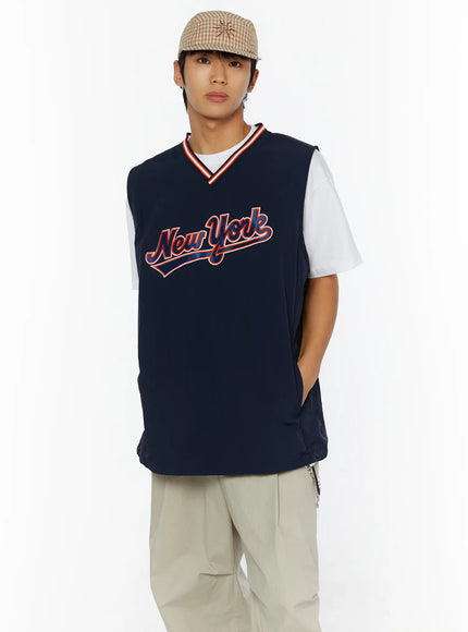 Men's Varsity Style Graphic Tank IL509 Dark blue close-up front shot on male model wearing plaid cap showing New York script graphic and red orange striped V-neck ribbed collar