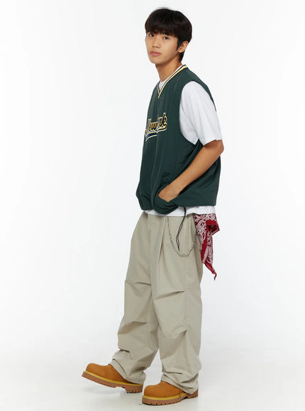 Men's Varsity Style Graphic Tank IL509 green side shot on male model with hands in pockets showing oversized silhouette layered over white tee with wide-leg trousers