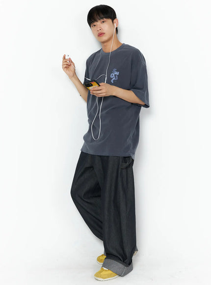 Men's Washed Wide Leg Jeans IL510 Black full-length front shot on male model leaning against wall in grey graphic tee holding MP3 player with earphones