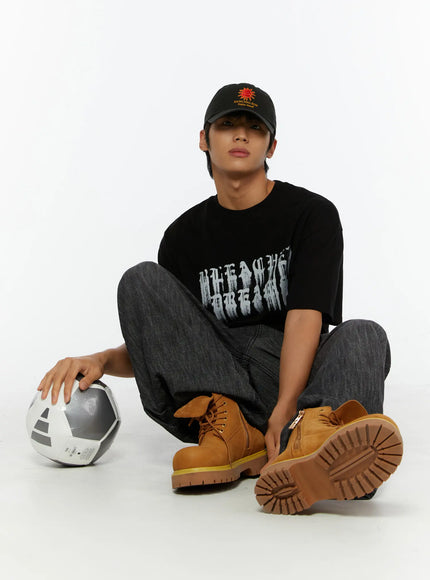 Men's Wide Leg Balloon Jeans IL515 Black lifestyle shot on seated male model holding soccer ball, styled with black graphic tee, black cap, and tan boots