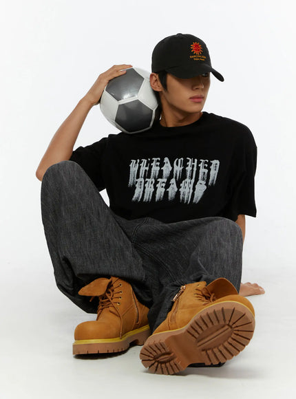 Men's Wide Leg Balloon Jeans IL515 Black lifestyle shot on seated male model holding soccer ball at shoulder, styled with black graphic tee, black cap, and tan boots