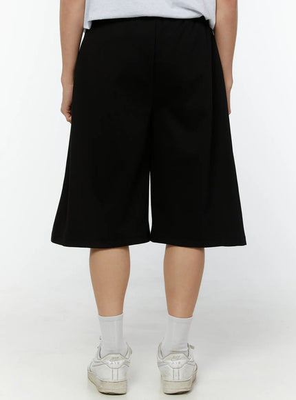 Men's Wide Leg Bermuda Sweat Shorts (Black) IU529 Black back view on model showing clean smooth fabric, elastic waistband, and wide-leg silhouette with white sneakers