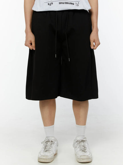LEWKIN Men's Wide Leg Bermuda Sweat Shorts (Black) IU529 Black front view on model wearing white graphic tee and sneakers, elastic drawstring waist, wide-leg silhouette