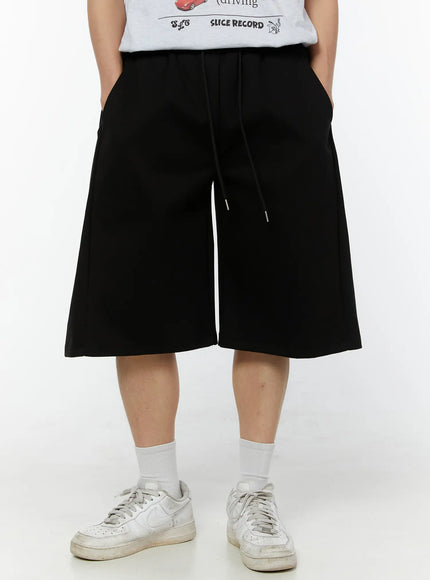 Men's Wide Leg Bermuda Sweat Shorts (Black) IU529 Black front view on model with hands in pockets, wide boxy cut, elastic waistband with drawstring, paired with white sneakers