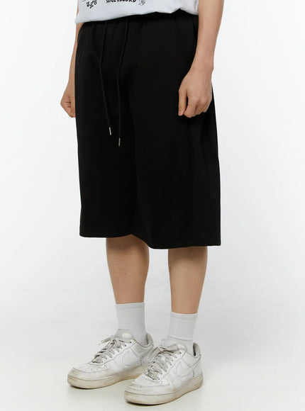 Men's Wide Leg Bermuda Sweat Shorts (Black) IU529 Black angled side view on model, drawstring detail visible, relaxed wide-leg fit styled with white sneakers