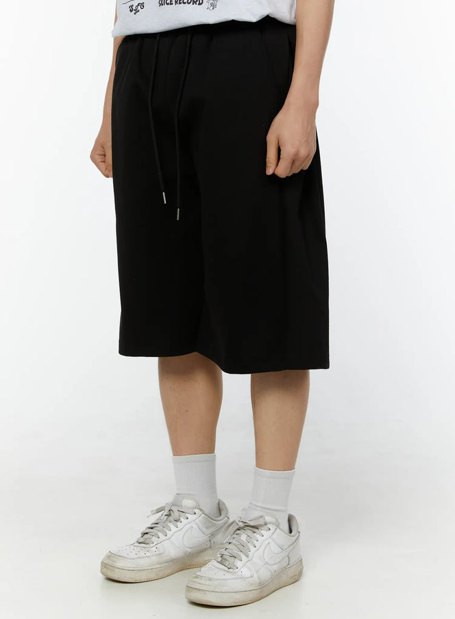 Men's Wide Leg Bermuda Sweat Shorts (Black) IU529 Black angled side view on model, drawstring detail visible, relaxed wide-leg fit styled with white sneakers