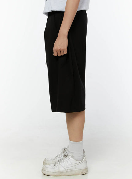 Men's Wide Leg Bermuda Sweat Shorts (Black) IU529 Black side view on model showing slim profile of wide-leg hem length, elastic waist with drawstring tip detail
