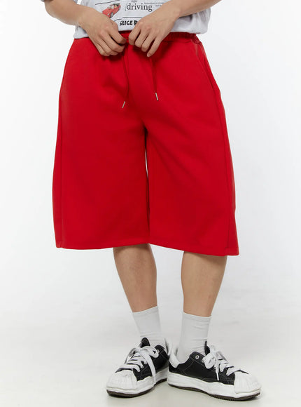Men's Wide Leg Bermuda Sweat Shorts (Red) IU529 Red front view on model standing straight, oversized wide-leg cut with pleated front and drawstring, paired with graphic tee and black sneakers