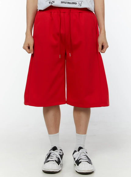 Men's Wide Leg Bermuda Sweat Shorts (Red) IU529 Red side view on model showing knee-length hem, side seam, and elastic waistband, styled with white socks and black sneakers