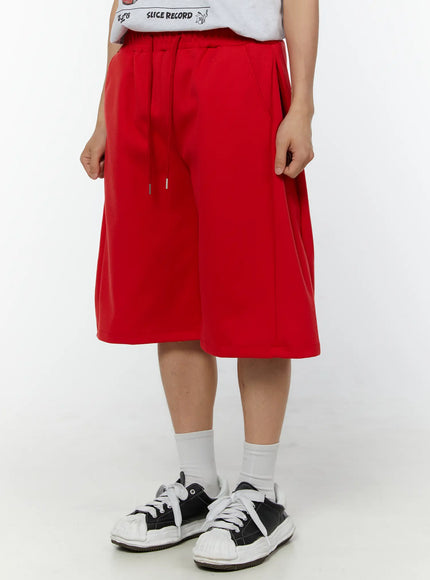 Men's Wide Leg Bermuda Sweat Shorts (Red) IU529 Red close-up side-front view on model with hands adjusting drawstring waistband, elastic waist and silver aglet detail