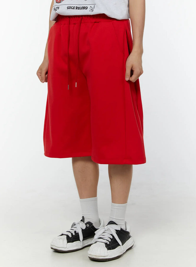 Men's Wide Leg Bermuda Sweat Shorts (Red) IU529 Red close-up side-front view on model with hands adjusting drawstring waistband, elastic waist and silver aglet detail