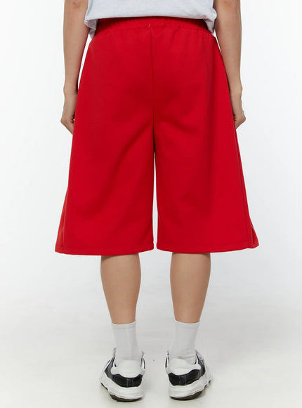Men's Wide Leg Bermuda Sweat Shorts (Red) IU529 Red close-up front view on model highlighting drawstring with silver metal aglets, pleated front panel, and elastic waistband texture