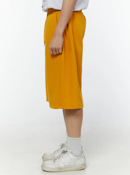 Men's Wide Leg Bermuda Sweat Shorts (Yellow) IU529 yellow side view on model revealing slim side profile, elastic waistband and knee-length hem with white sneakers
