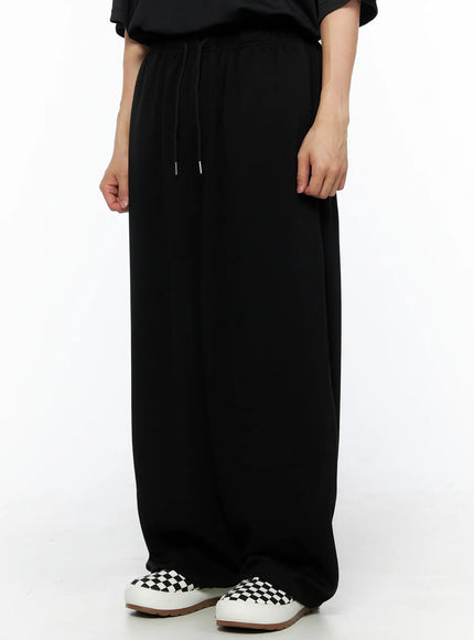 Men's Wide Leg Sweatpants (Black) IL514 black side-angle view on model showing wide-leg drape, drawstring waist, and cargo pocket detail styled with black tee and checkered sneakers
