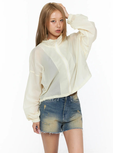 Nylon Cropped Windbreaker CU523 Light beige front model shot, hand raised to hair, sheer translucent balloon-sleeve jacket, mock neck, cropped hem, paired with distressed denim shorts