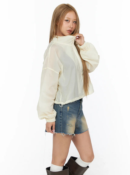Nylon Cropped Windbreaker CU523 Light beige side model shot, close three-quarter view, sheer semi-transparent fabric, zip detail at collar, drawstring hem, worn with denim shorts