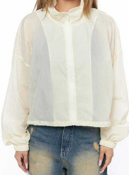 Nylon Cropped Windbreaker CU523 Light beige close-up front detail shot, sheer mesh texture panel, center white zipper, elastic cropped hem, and semi-transparent grid fabric visible