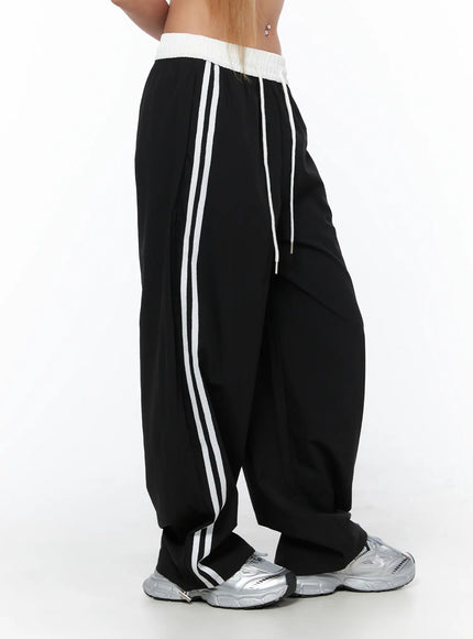 Nylon Track Pants CU523 Black side close-up on model showing white ribbed waistband, double white stripe detail, and relaxed wide-leg cut