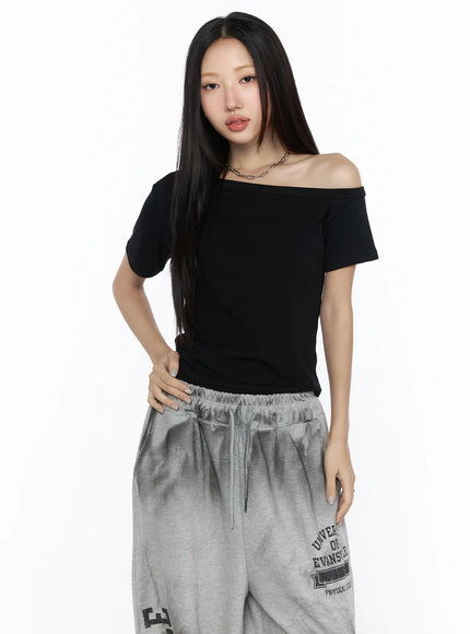 Off-Shoulder Casual Tee CL527 Black three-quarter front shot on model hand on hip, asymmetric off-shoulder neckline, fitted crop silhouette, paired with grey drawstring sweatpants