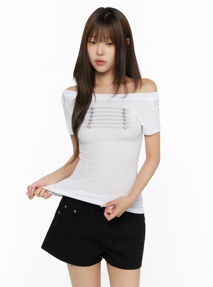 Off-Shoulder Crop Top IL523 White front shot on model pulling hem outward, showcasing cropped length and silver cord button detail on off-shoulder ribbed knit top
