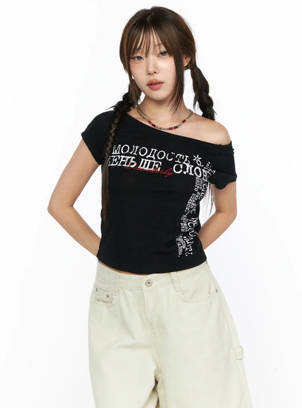 Off-Shoulder Graphic Crop Short Sleeve Tee CL514 Black front view on model with one-shoulder styling Russian Cyrillic graphic print paired with beige shorts