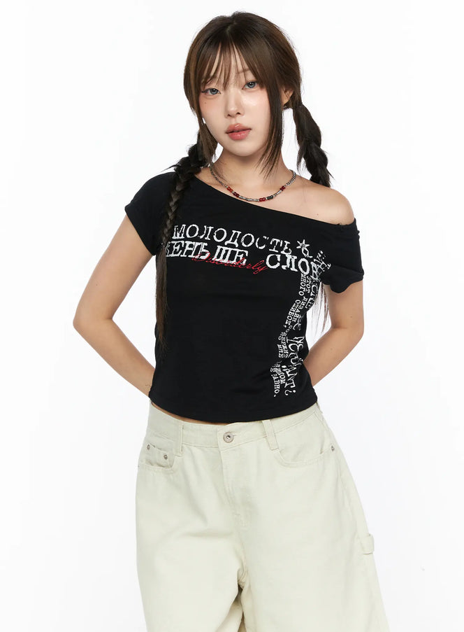 Off-Shoulder Graphic Crop Short Sleeve Tee CL514 Black front view on model with one-shoulder styling Russian Cyrillic graphic print paired with beige shorts