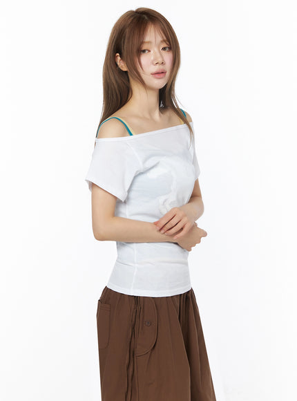 Off-Shoulder Graphic Short Sleeve Top IU525 White side shot on model with arms crossed, fitted silhouette, teal and beige bra straps layered over off-shoulder neckline