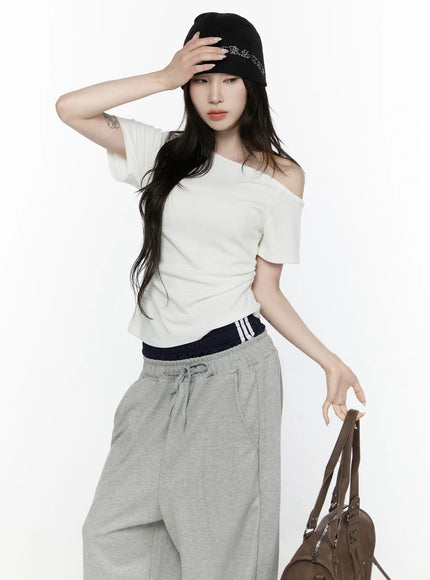 Off-Shoulder T-Shirt IL522 White front shot on model holding hat, asymmetric shoulder drop, ruched waist detail, styled with grey joggers