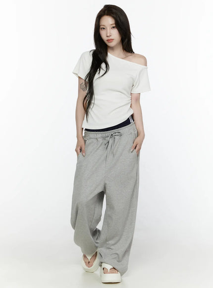 Off-Shoulder T-Shirt IL522 White full-body front shot on model standing straight, one-shoulder silhouette, hands at sides, styled with grey joggers