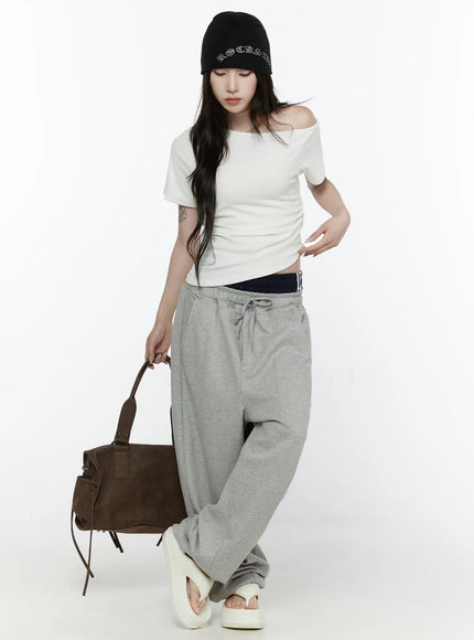 Off-Shoulder T-Shirt IL522 White full-body shot on model looking down, asymmetric neckline, styled with grey sweatpants and sandals