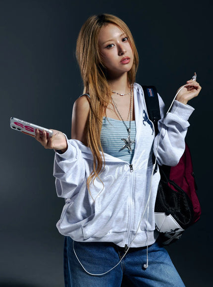 Oversized Graphic Zip-Up Hoodie CL528 Light Gray lifestyle three-quarter front shot on model in dark studio holding phone with backpack wearing open hoodie over striped cami and jeans
