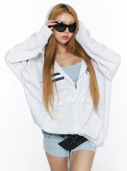 Oversized Graphic Zip-Up Hoodie CL528 Light Gray front close-up on model pulling up hood with both hands showing Saddle script graphic and navy horse emblem on chest