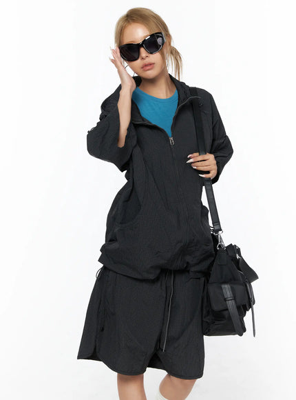 Oversized Plaid Windbreaker CU523 Black styled front shot model wearing zip-up windbreaker with black crossbody bag sunglasses teal top matching shorts hand raised near face