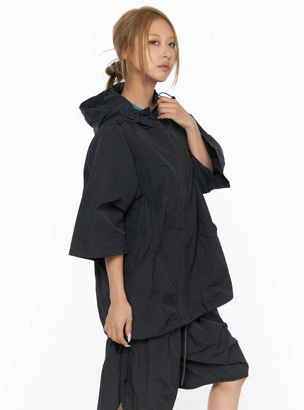 Oversized Plaid Windbreaker CU523 Black close-up side shot model holding drawstring hood detail showing grid-check nylon texture zip closure and oversized sleeve with teal top visible