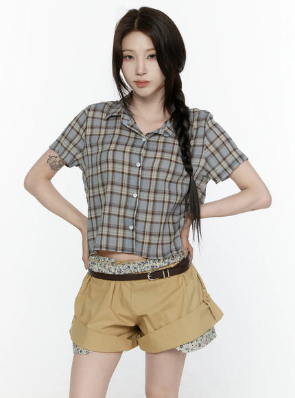 Plaid Button-Up Shirt IL521 Blue front shot on model with braided hair hands on hips, cropped blue-grey plaid short-sleeve shirt with open collar and white buttons, layered over floral top with khaki shorts