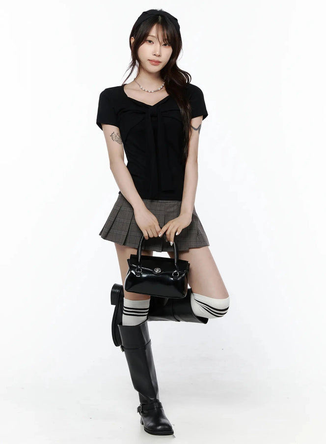 Plaid Pleated Mini Skirt IL506 Brown full-body front shot on model holding handbag one knee raised styled with black bow top striped knee socks and tall buckle boots