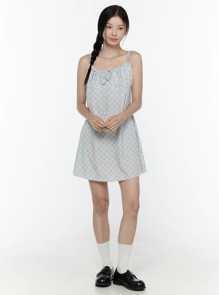 Plaid Shirred Cami Mini Dress IL506 Light blue front model shot no accessories model holding tie cord showing diagonal plaid print and gathered bust detail