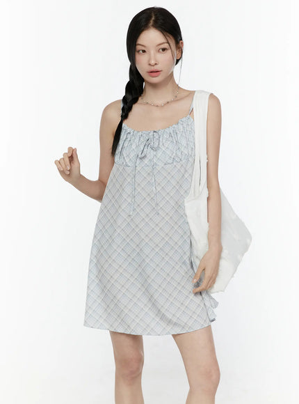 Plaid Shirred Cami Mini Dress IL506 Light blue three-quarter front model shot carrying white tote bag hand raised showing ruched bust and loose mini silhouette
