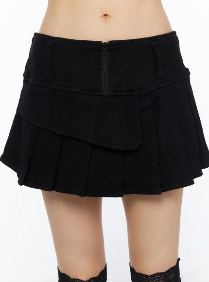 Pleated Mini Skirt CU530 Black close-up front detail showing asymmetric layered flap, center back zipper, and pleated hem on model