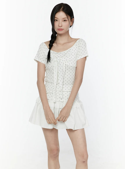 Polka Dot Round Shoulder Top IL506 White front model shot hands folded, wide scoop neckline, black polka dot fabric, center tie ribbon detail and fitted waist with white layered ruffle skirt