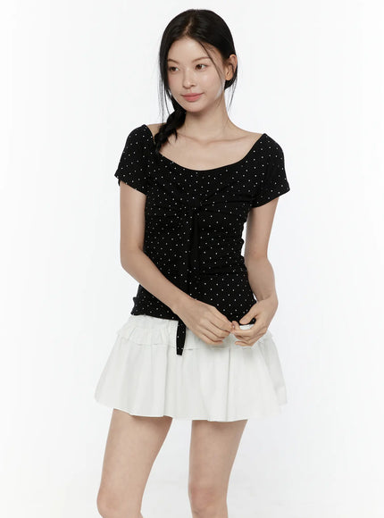 Polka Dot Round Shoulder Top IL506 Black front model shot, white polka dot pattern on black fabric, scoop neckline, center tie ribbon, fitted short-sleeve top styled with white tiered ruffle mini skirt