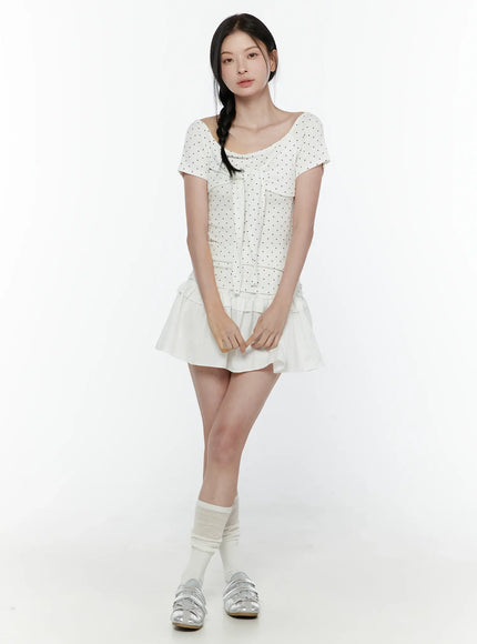 Polka Dot Round Shoulder Top IL506 White full-length front model shot hands clasped, black polka dot tie-front top with scoop neckline paired with white ruffle skirt, white socks and silver sandals