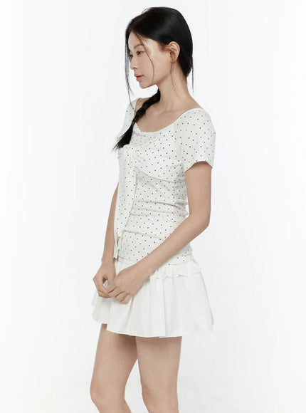 Polka Dot Round Shoulder Top IL506 White side model shot showing fitted tie-front bodice with ruched waist, black polka dot pattern, short sleeves and layered white ruffle skirt hem