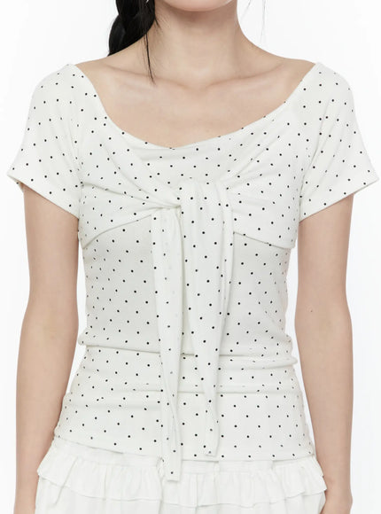Polka Dot Round Shoulder Top IL506 White close-up front model shot highlighting scoop neckline, center tie ribbon detail, ruched bust seaming and black polka dot knit fabric texture