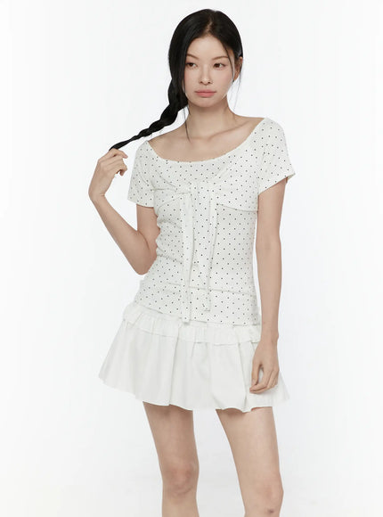 Polka Dot Round Shoulder Top IL506 White front model shot with model touching braid, wide scoop neckline, black polka dot tie-front top styled with white tiered ruffle mini skirt, no accessories