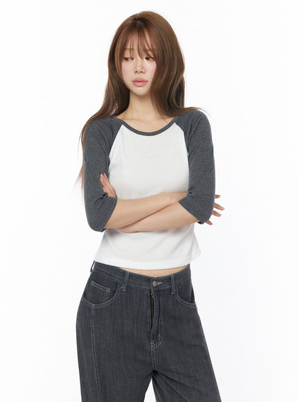 Raglan Shirred 3/4 Top IU525 White front on model with long hair, arms crossed, showcasing cropped hem and charcoal raglan sleeve contrast, styled with dark wide-leg denim