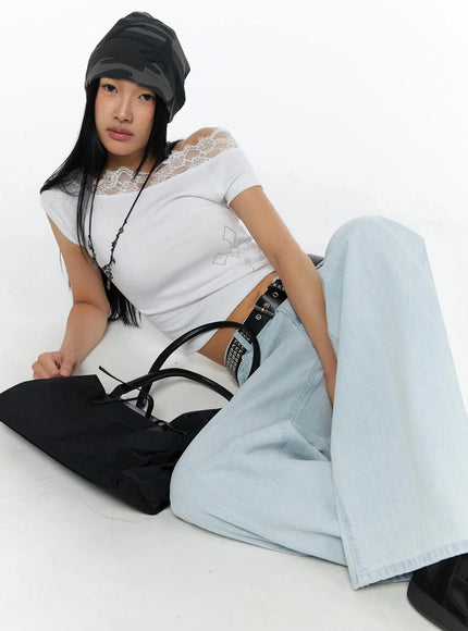 Rhinestone Cross Lace Trim Off-Shoulder Top IL516 White lifestyle front shot model seated on floor wearing camo beanie cross necklace studded belt holding black bag styled with light blue wide-leg jeans