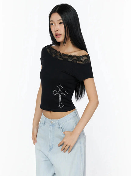 Rhinestone Cross Lace Trim Off-Shoulder Top IL516 Black three-quarter front shot model standing wearing fitted crop top with lace neckline and rhinestone cross motif styled with light blue jeans